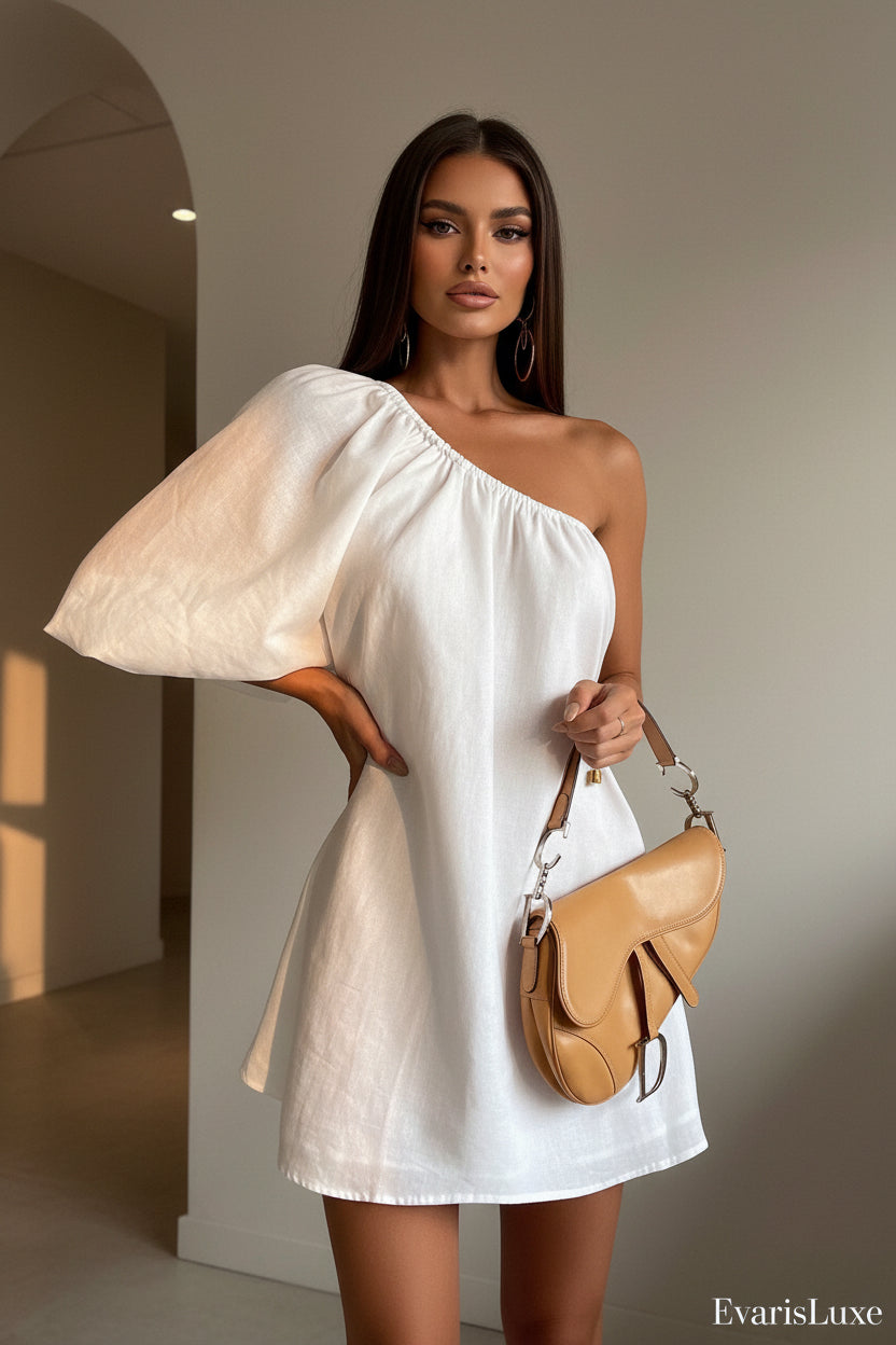 Woman wearing a white one-shoulder dress holding a tan handbag against a plain background