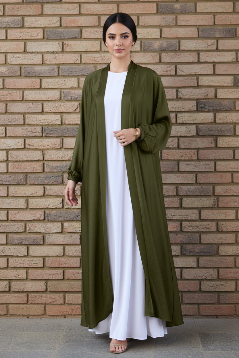 Person wearing an olive green abaya over a white outfit against a brick wall.