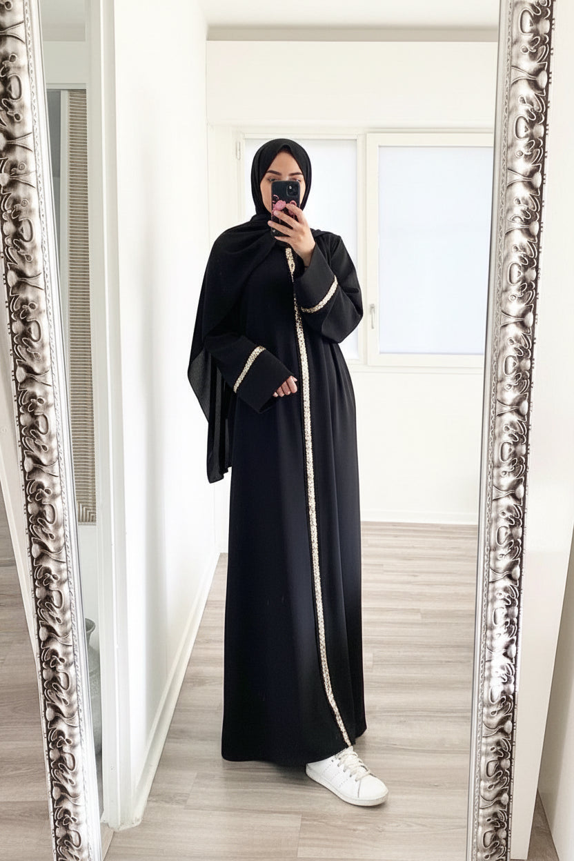 Person wearing a black abaya with white trim in front of a decorative mirror.