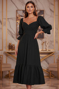 Black dress with puffed sleeves worn by a person standing against a white wall with green foliage.