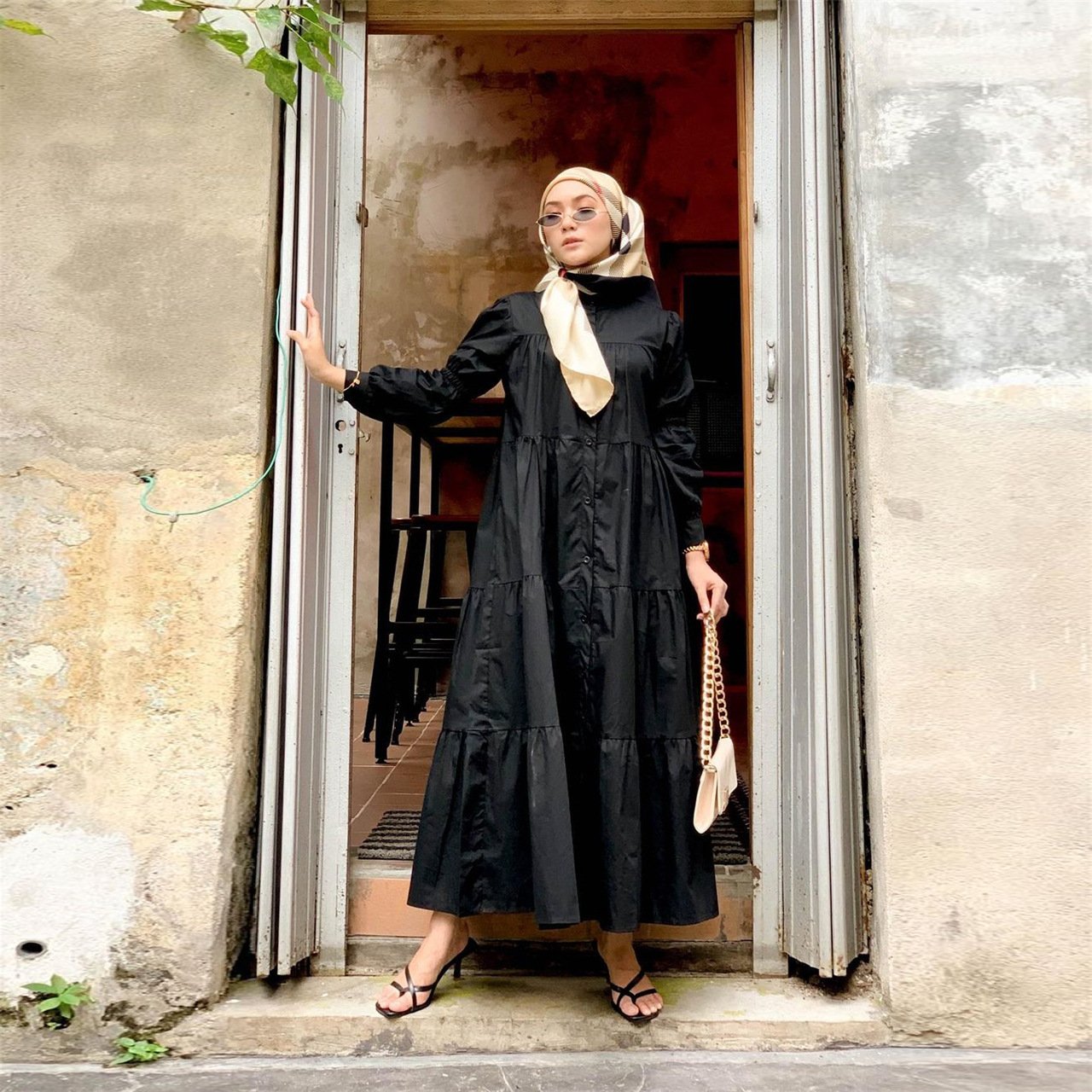 Woman in a black dress and beige hijab standing in an open doorway.