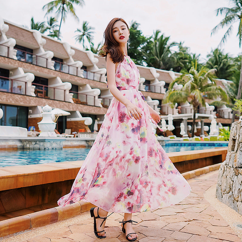 Woman in a pink floral dress standing by a pool with resort buildings and palm trees in the background.