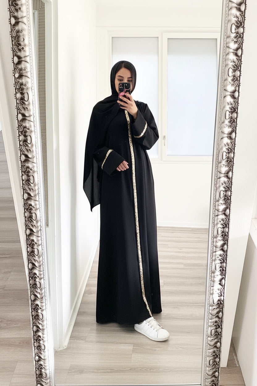 Person wearing a black abaya with white trim in front of a decorative mirror.