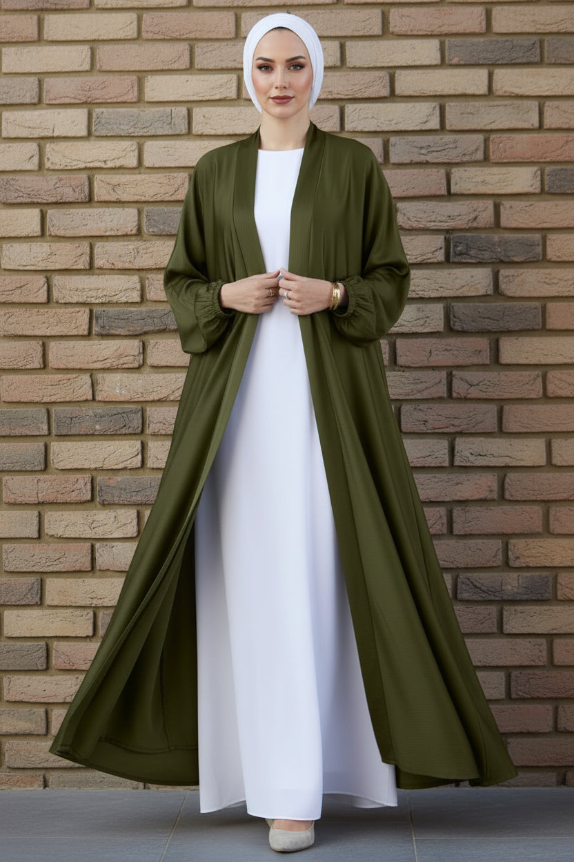 Person wearing an olive green abaya over a white outfit against a brick wall.