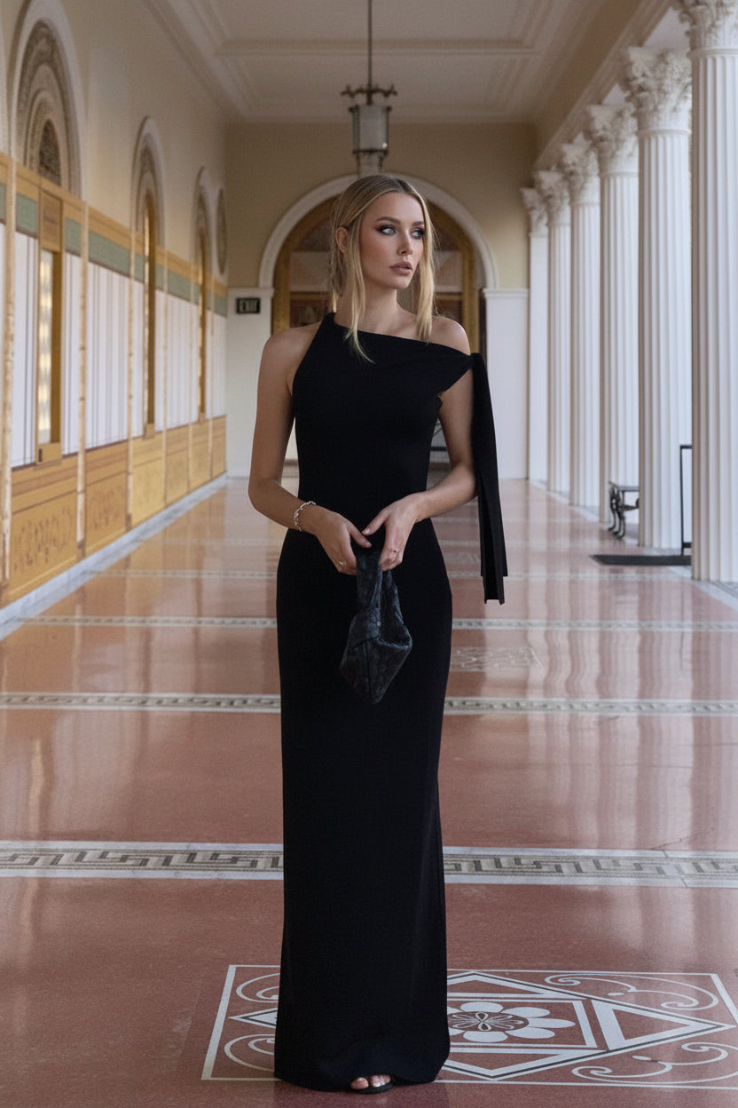 Woman in a black one-shoulder evening gown holding a clutch in an elegant hallway.