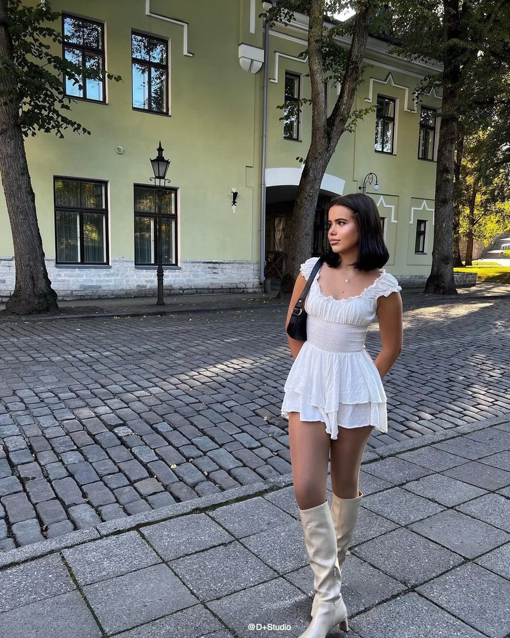 Woman in a white dress and beige boots walking on a street with a building in the background