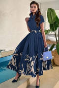 Woman in a blue dress with a floral pattern holding a blue handbag by a pool.
